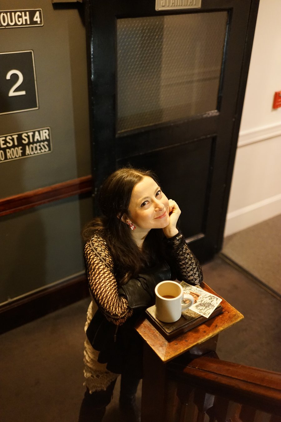 Jay Lynn resting her chin on one hand beside a cup of coffee on a small wooden table, in a dark stairwell lit by warm light