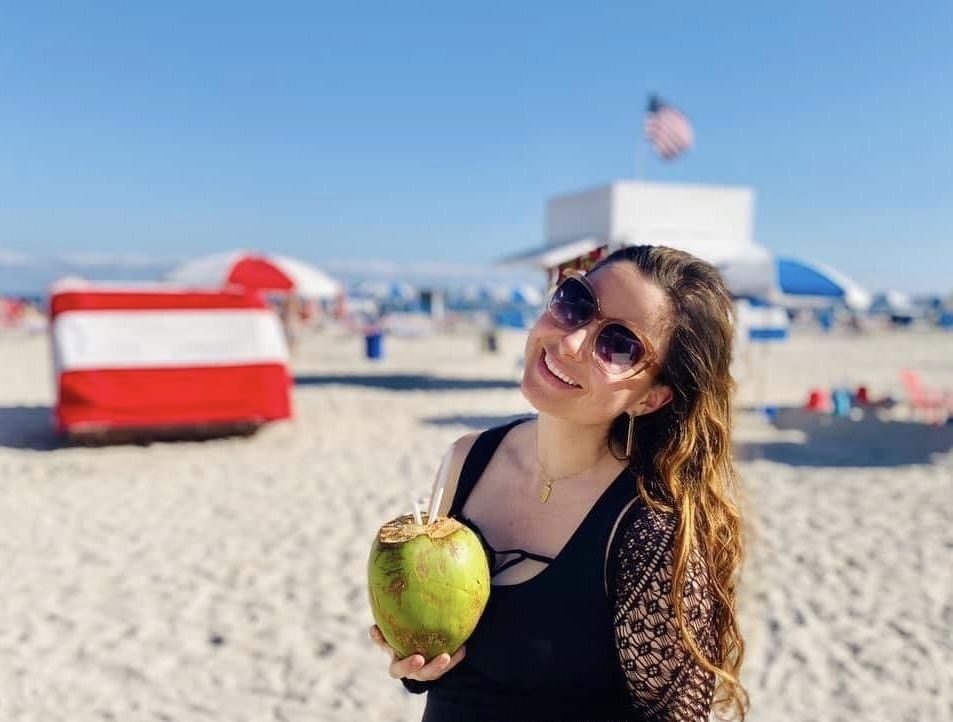 Jay Lynn on a beach in sunglasses, holding a green coconut with a straw in it, beach umbrellas and a lifeguard hut behind
