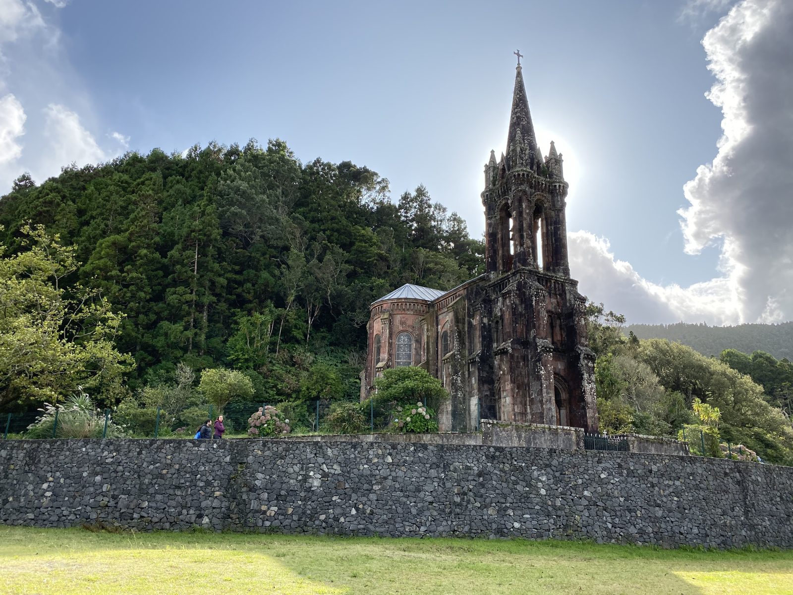 A gothic revival chapel with a tall stone spire, backlit, against a wooded hillside