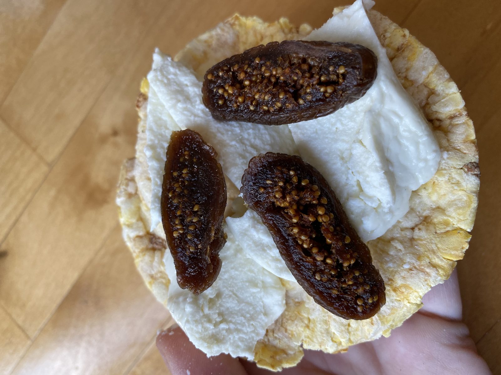 A hand holding a rice cake topped with fresh white cheese and three dried figs