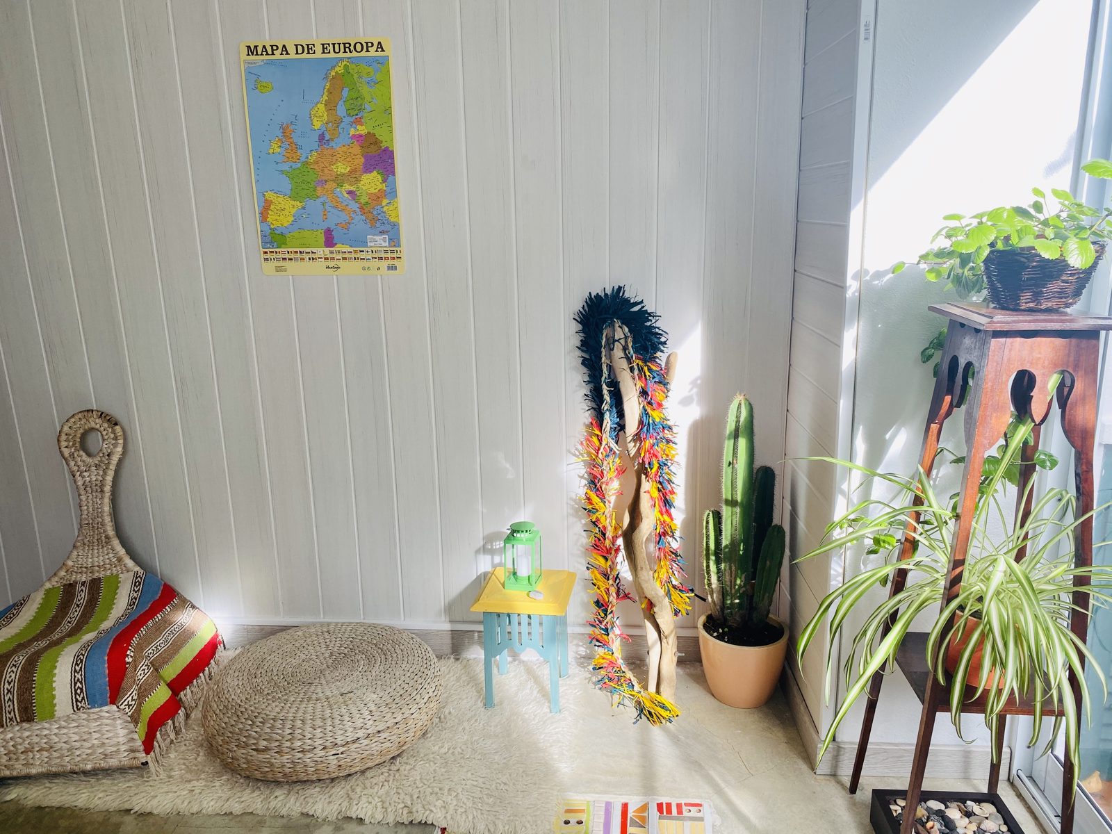 The corner of a rental with a map of Europe on the wall, plants, a woven floor cushion and a small blue table