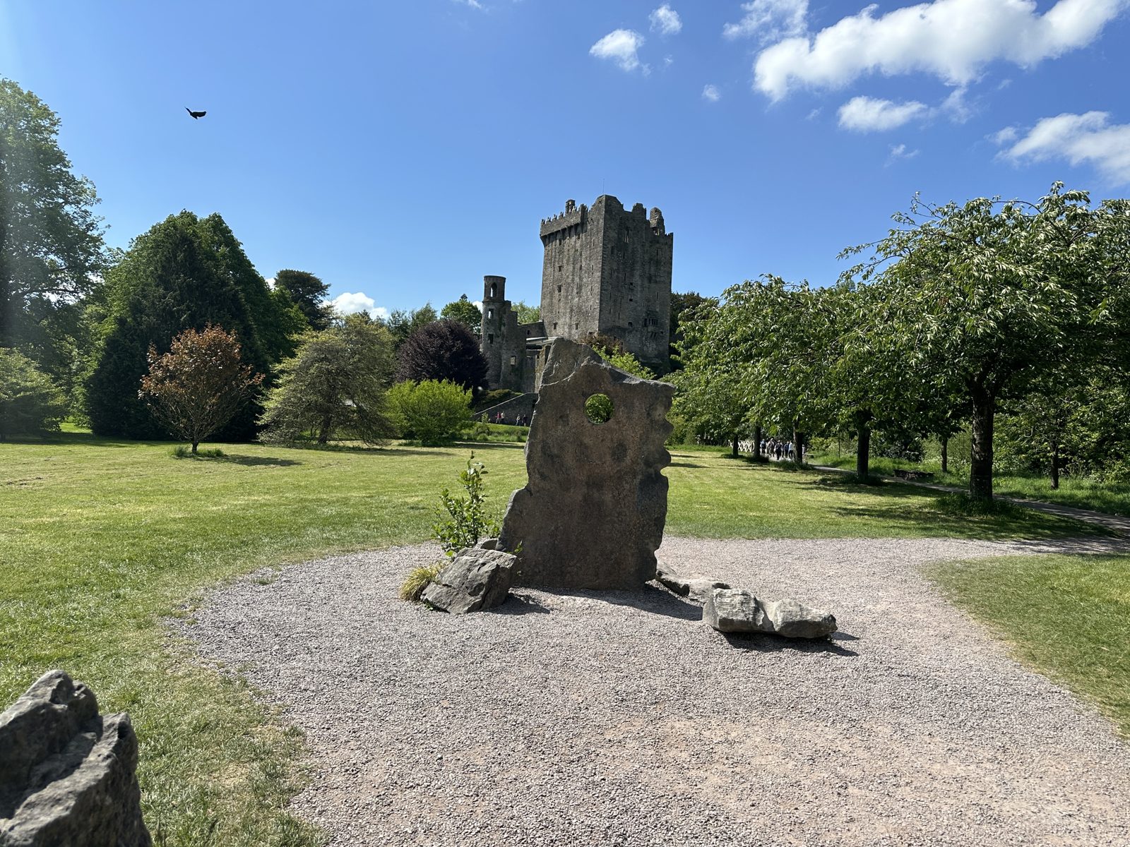 Blarney Castle seen across a sunlit lawn, with a tall pierced standing stone in the foreground