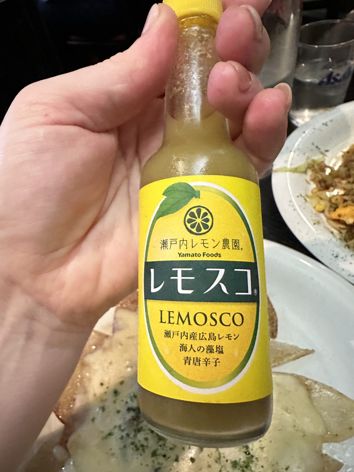 A hand holding a small yellow bottle of Lemosco lemon chilli sauce over a plate of okonomiyaki on a griddle