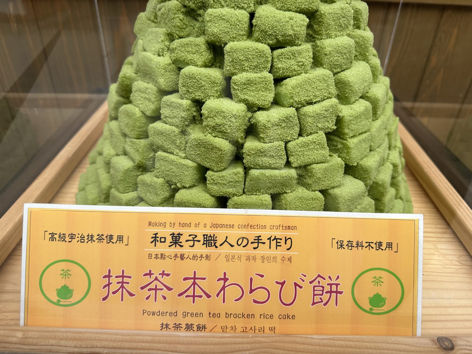 A pyramid of green matcha dusted warabi mochi cubes in a wooden tray behind glass, with a handwritten sign