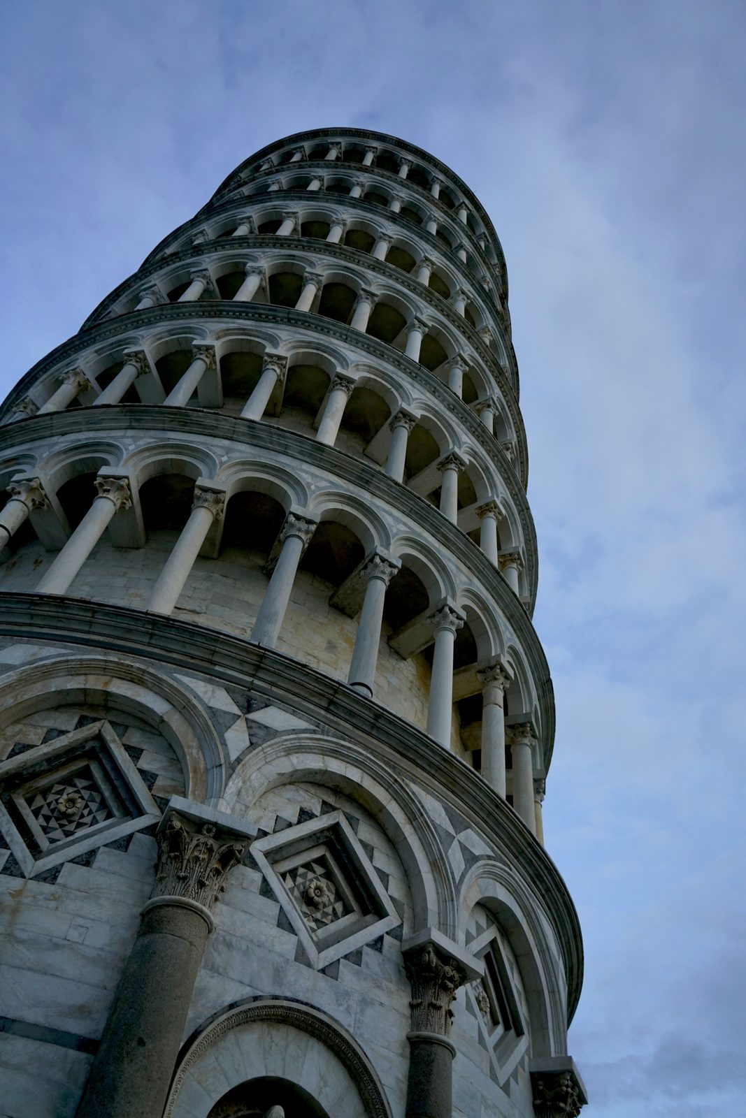 The Leaning Tower of Pisa shot from directly below, its tiers of white marble columns curving away against a pale blue sky