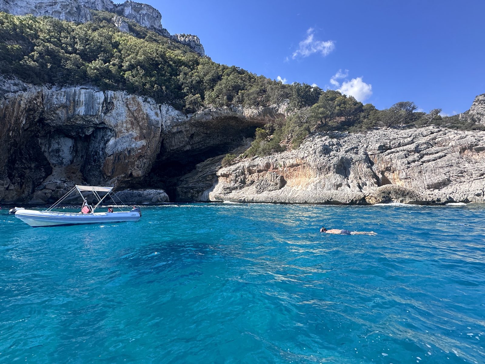 Bright turquoise water in the Gulf of Orosei below white limestone cliffs, with a small boat moored beside a cave mouth and one person snorkelling