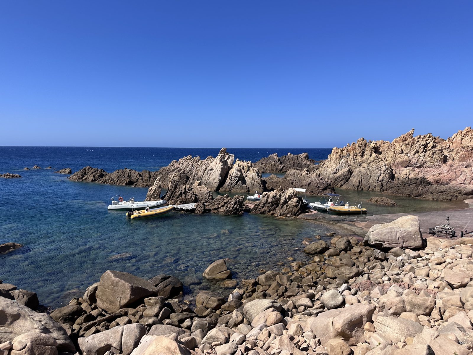 A cove on the north coast with jagged pink granite stacks, inflatable boats moored in clear shallow water and a rocky shoreline in the foreground