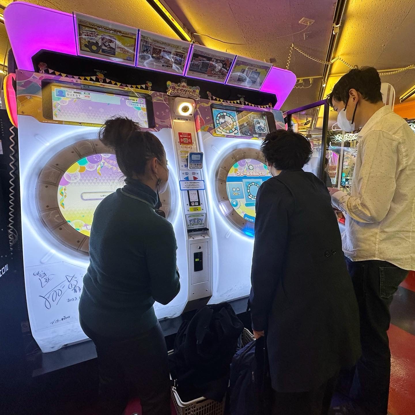 Three people standing at a brightly lit rhythm arcade machine in a Japanese game centre