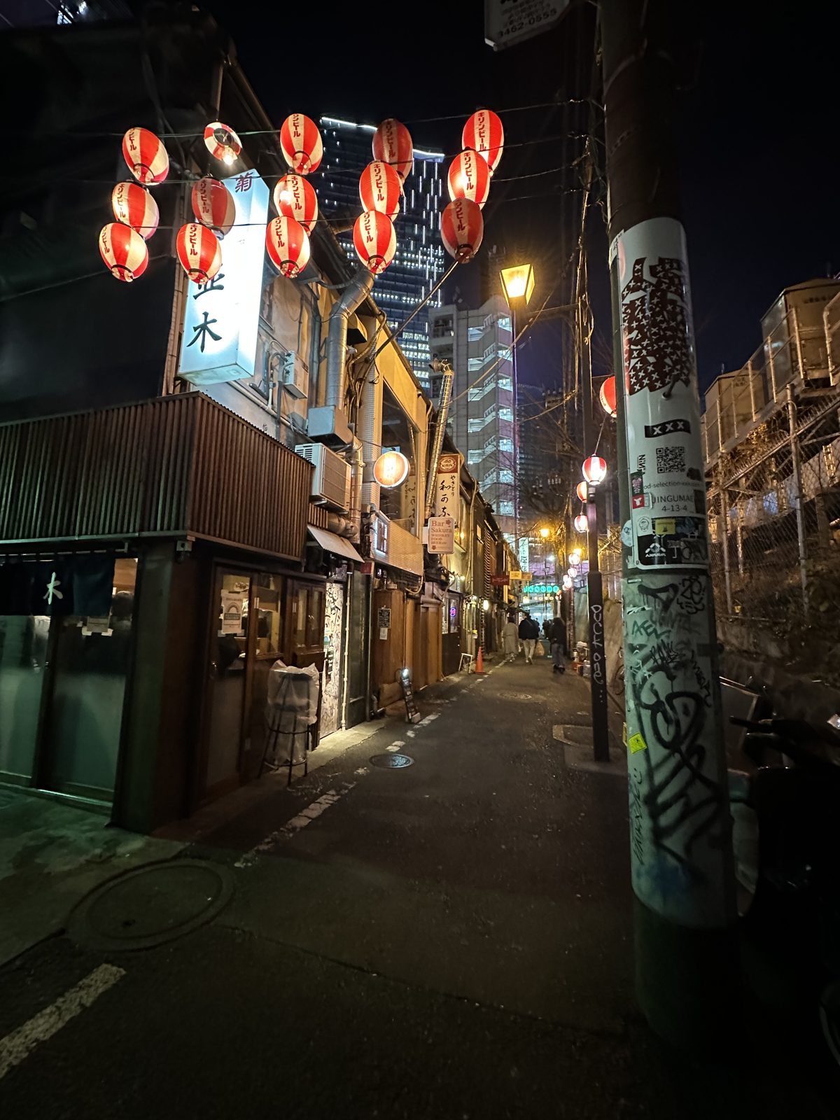 A narrow alley at night lined with tiny bar fronts and red paper lanterns, under a railway overpass