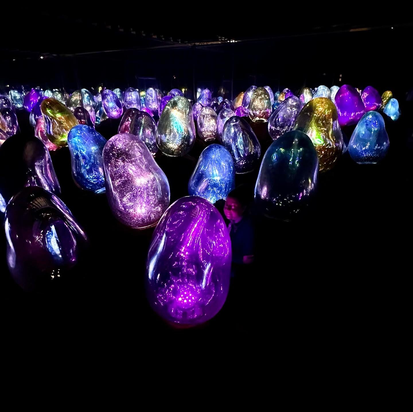 Dozens of large glowing ovoids in a dark space, lit purple, blue and gold, with someone standing among them