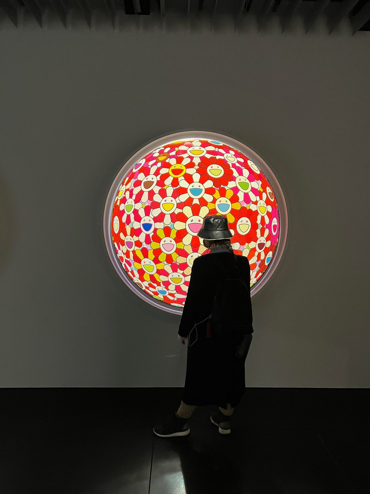 A visitor in front of a circular Takashi Murakami panel of coloured flowers, lit from behind in a dark room