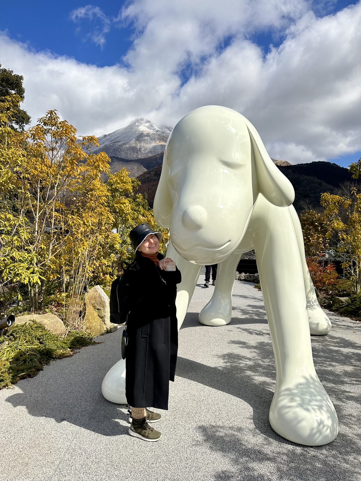 A visitor beside Yoshitomo Nara's giant white dog sculpture on the Comico Art Museum terrace in Yufuin, with snow on Mount Yufu behind