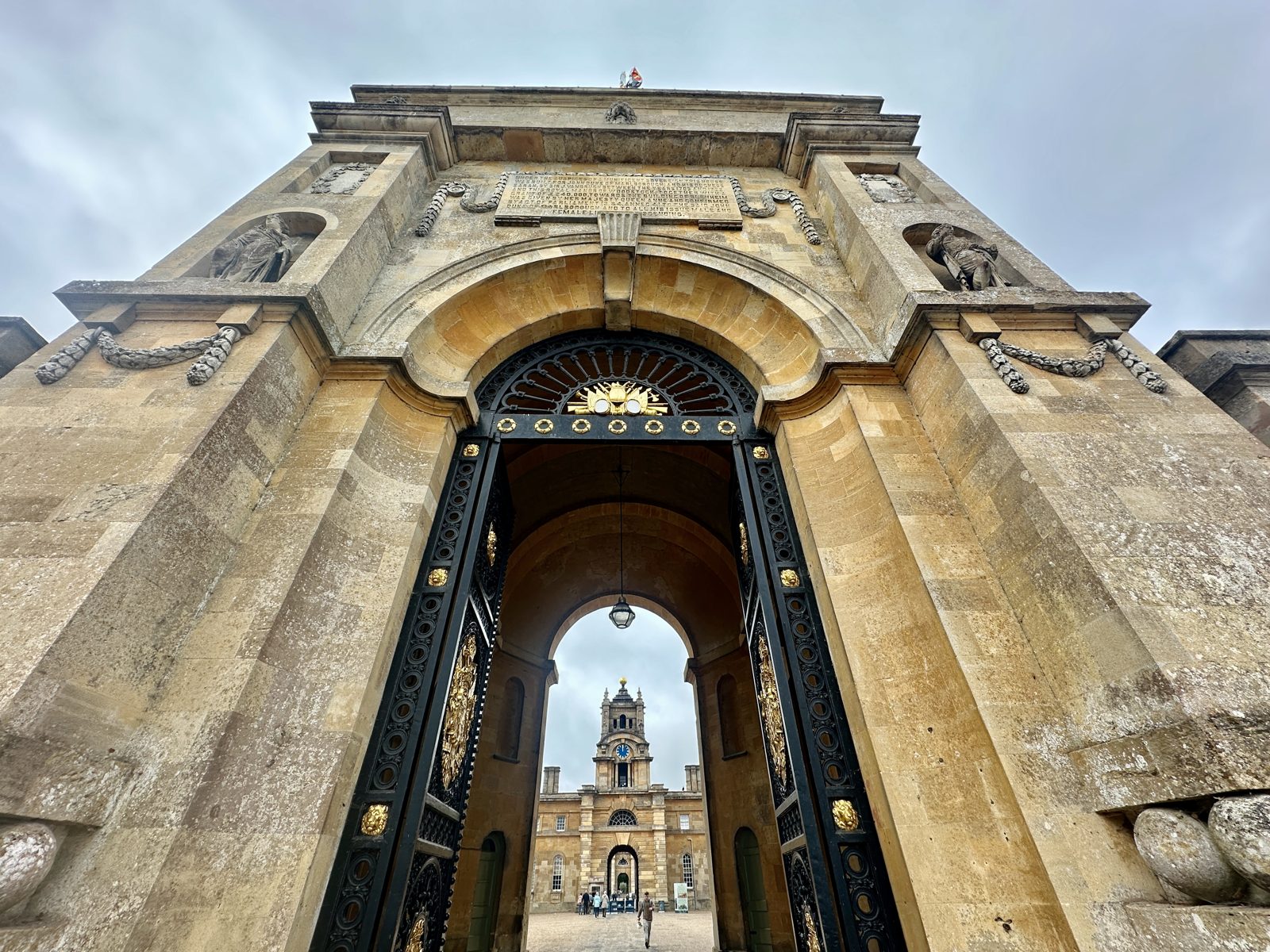 The triumphal arch at Blenheim Palace seen from below, its tall gilded iron gates standing open with the clock tower of the Great Court framed in the opening
