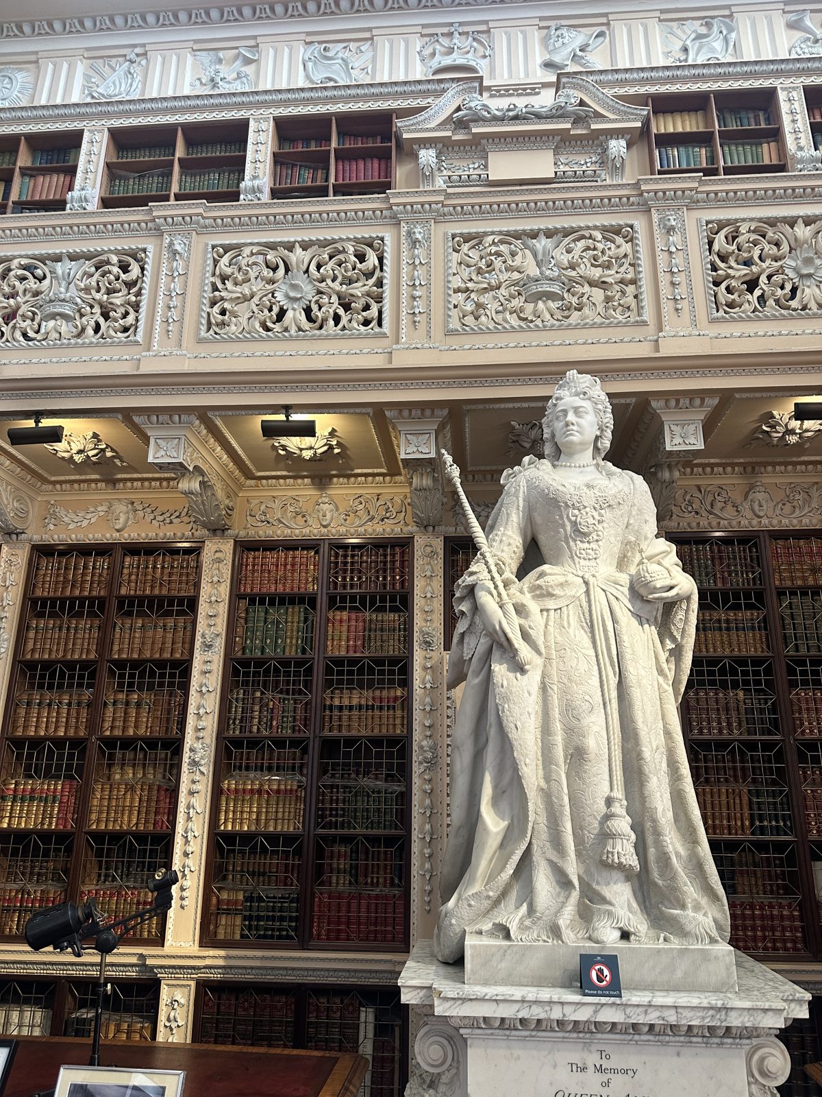 A marble statue of Queen Anne holding a sceptre, standing in front of tall glazed bookcases in the Long Library at Blenheim, under heavy carved white plasterwork