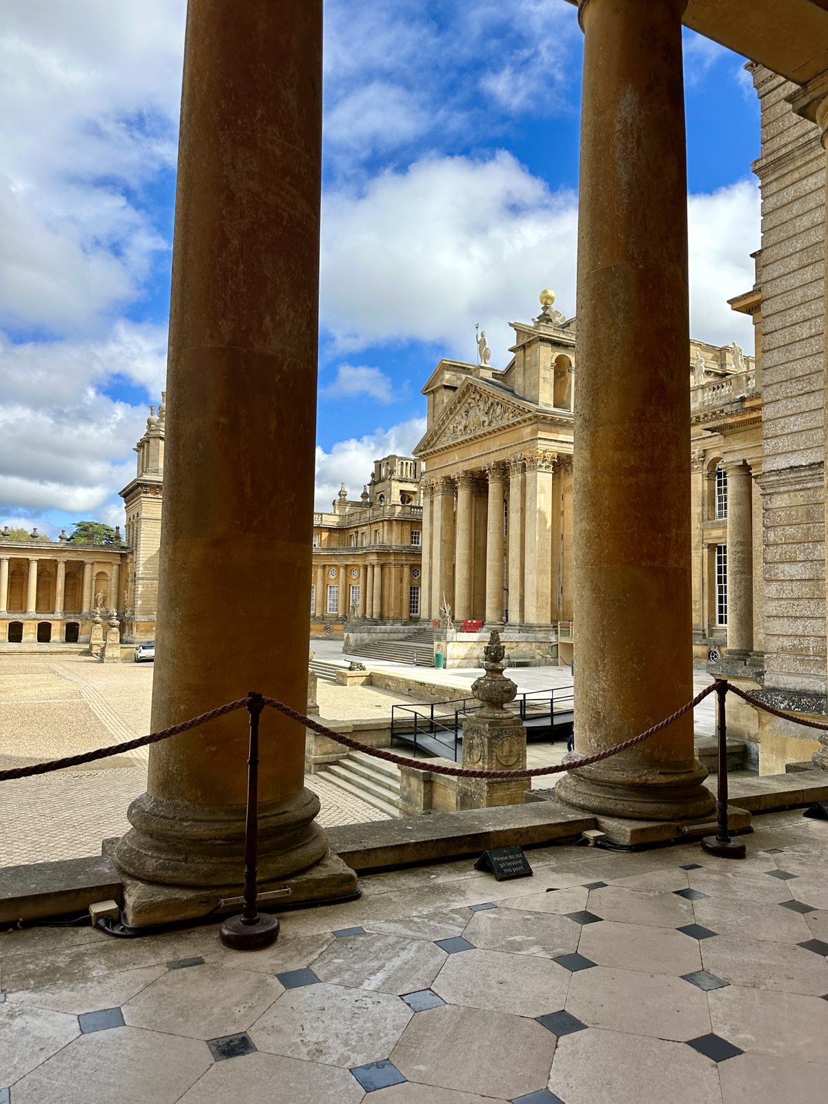 Looking out between two tall stone columns of the palace portico, across the Great Court to the far wing with its carved pediment and a gilded finial against a blue sky