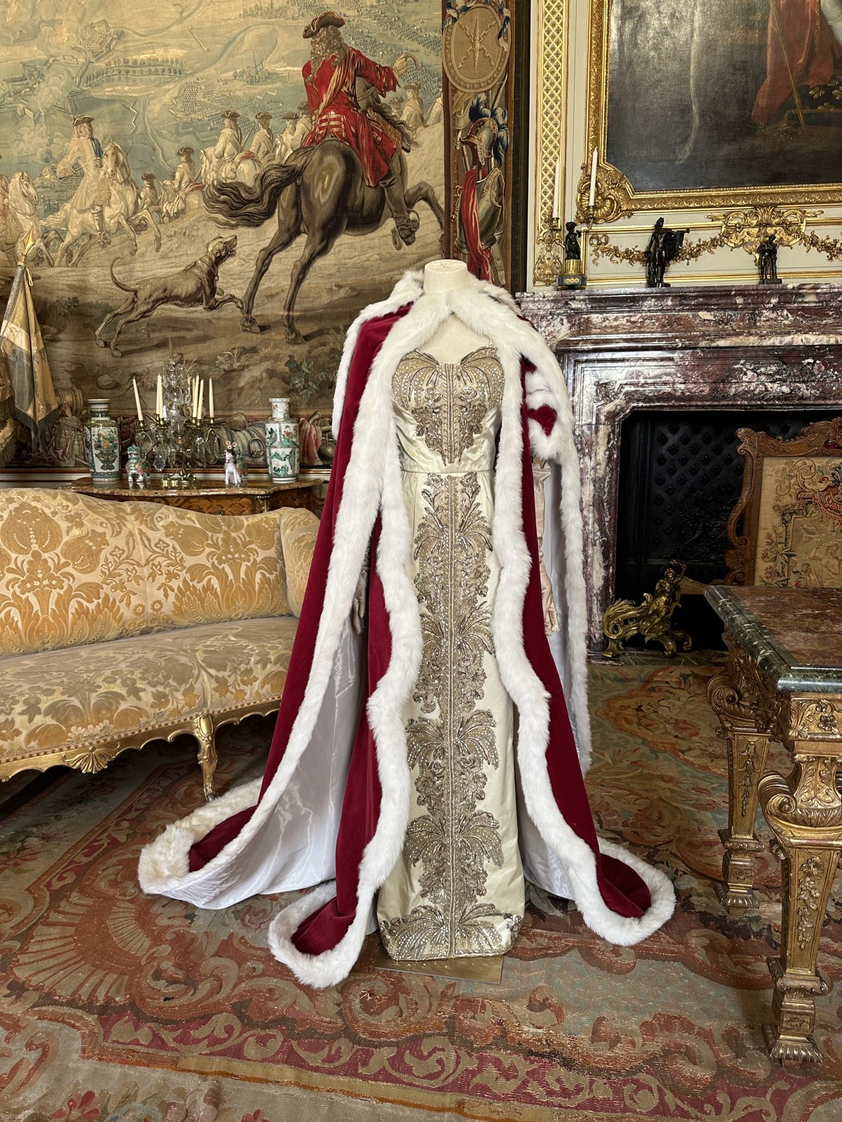 A coronation robe of crimson velvet trimmed with white fur and heavy silver embroidery, displayed in a room hung with tapestries