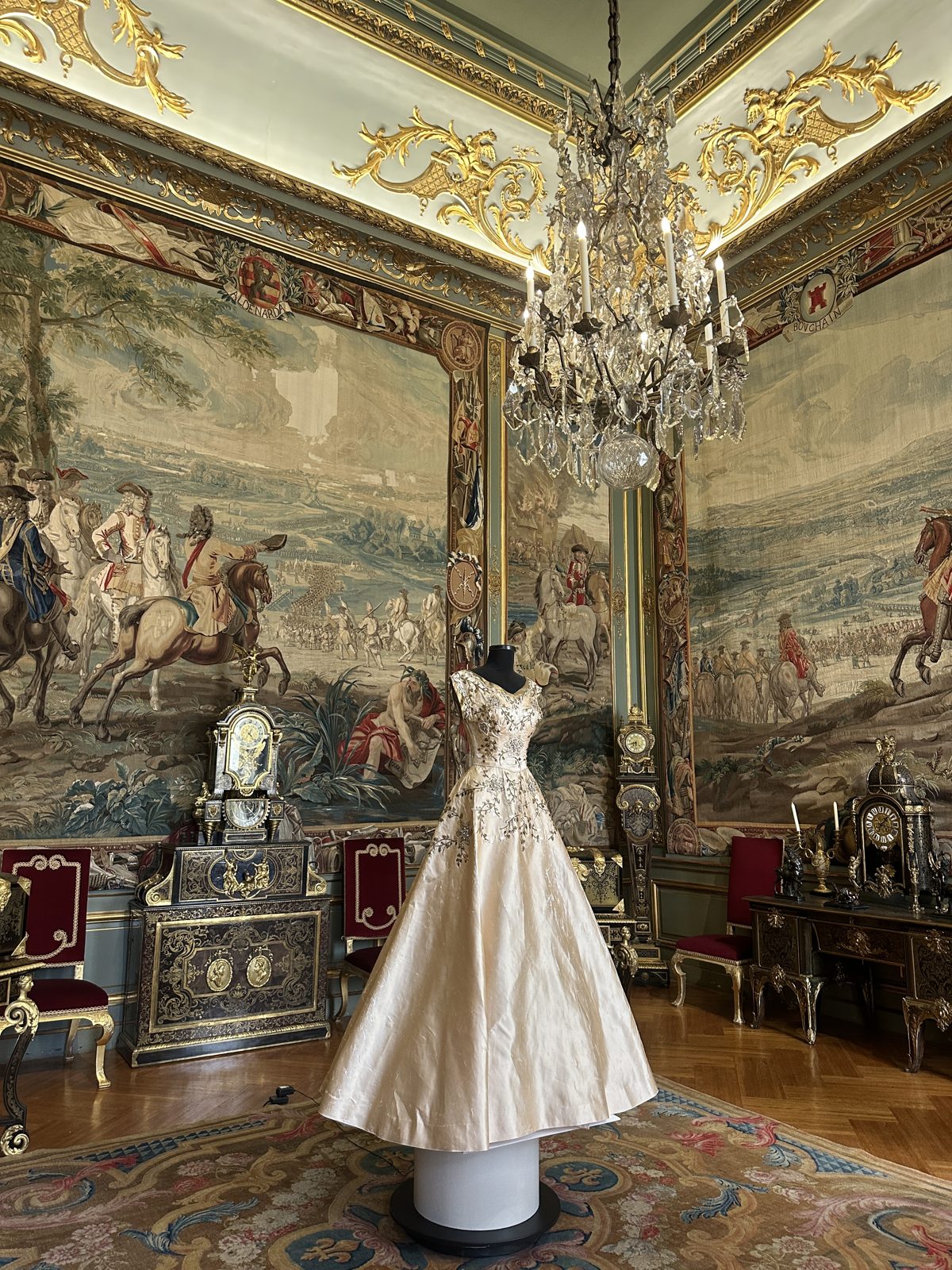 A state room at Blenheim hung with large tapestries of mounted soldiers, a crystal chandelier overhead and a pale embroidered gown on a stand