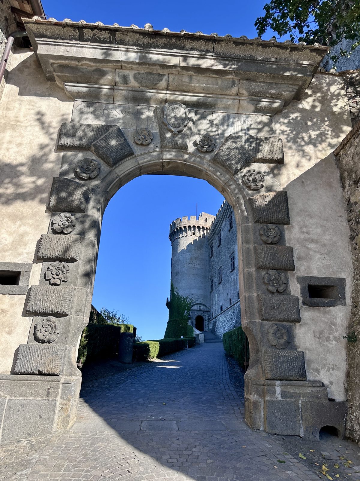 A carved stone archway in Bracciano framing the round tower of Castello Orsini-Odescalchi, with a stone paved lane and clipped hedges leading up to the castle entrance