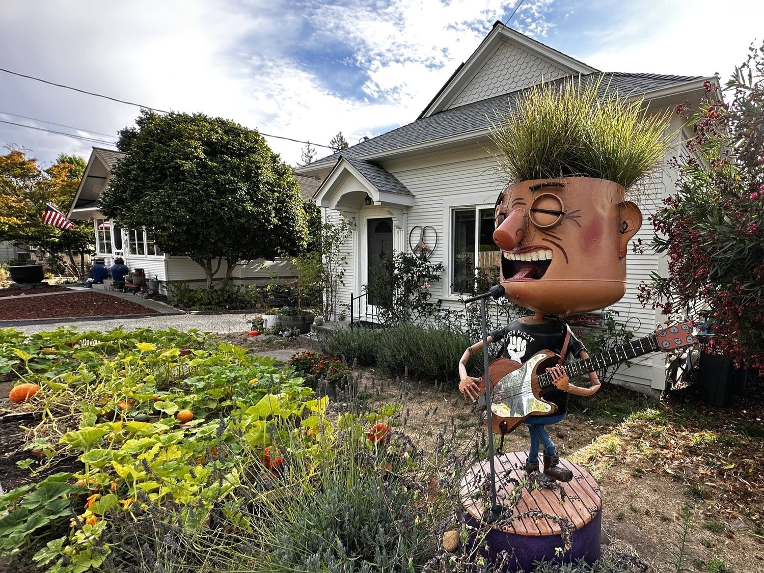 A junk art musician with a flowerpot head sprouting grass, playing guitar at a microphone in front of a white cottage and pumpkin patch