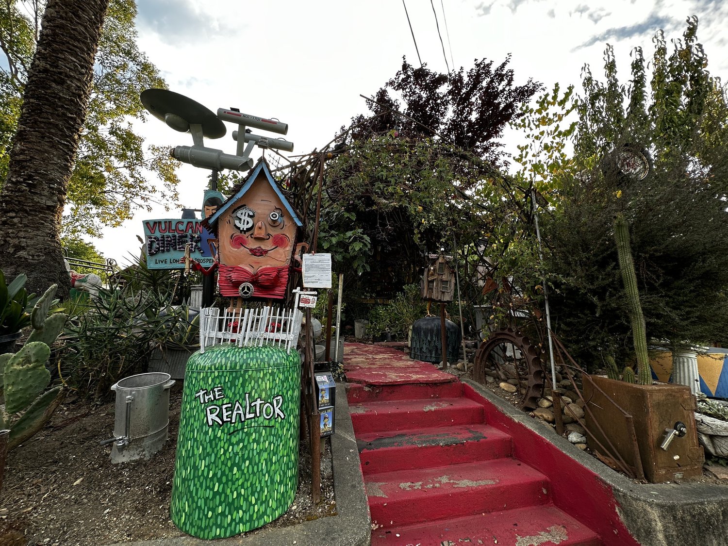 The Realtor sculpture, a house headed figure with a dollar sign eye, beside red painted steps and a Vulcan Diner sign