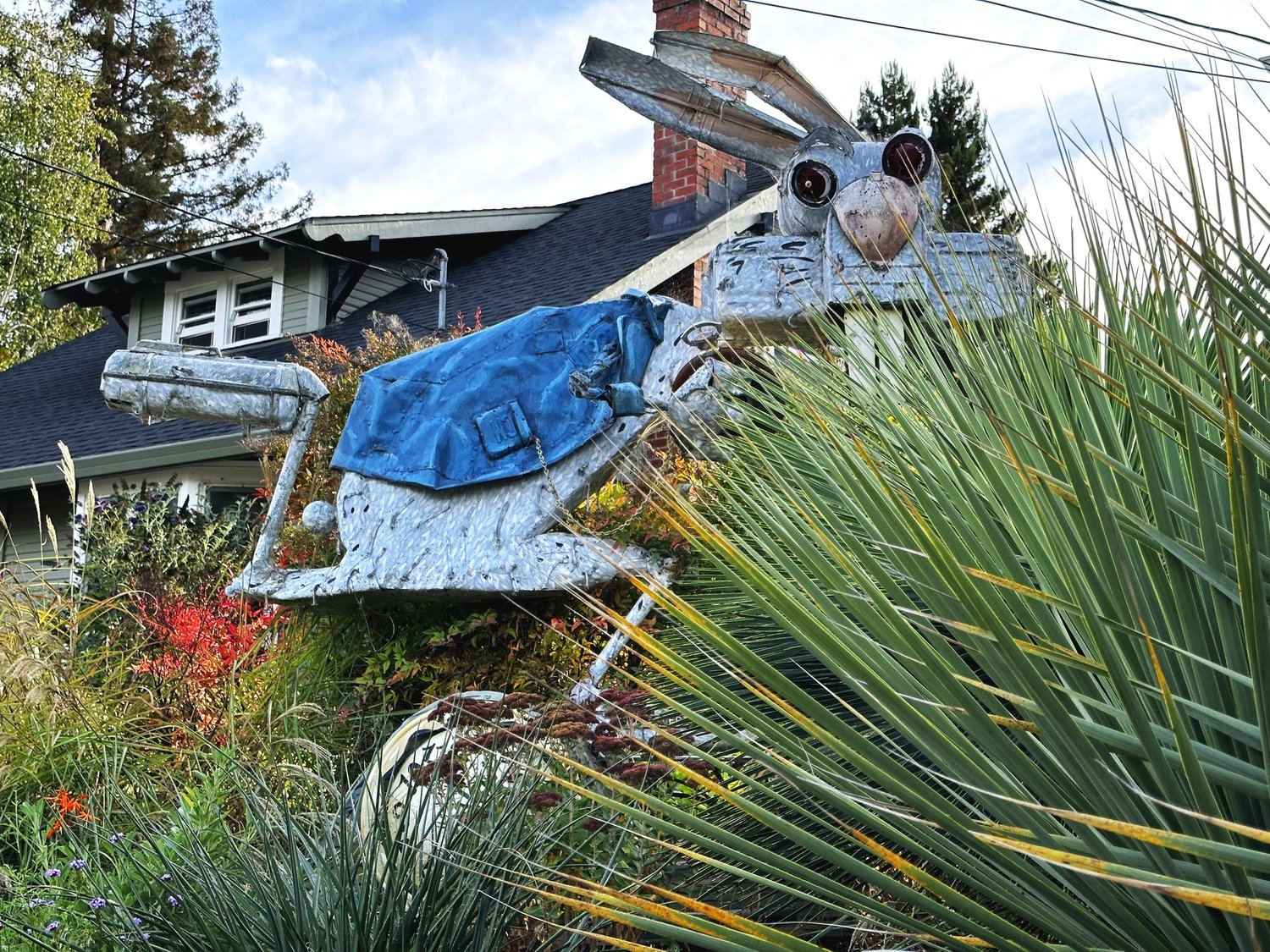 A galvanised metal hare sculpture wearing a blue saddle blanket, leaping above a garden of grasses