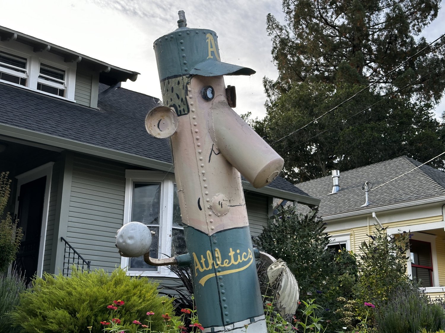A tall Oakland Athletics baseball player built from scrap metal drums, holding a ball in a front garden