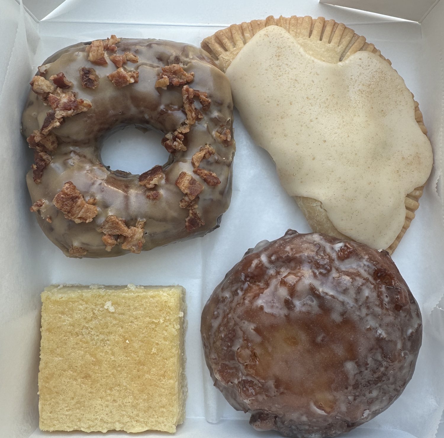A box of gluten free bakes from Blue Moon Bakery: a glazed doughnut topped with bacon, a hand pie, a lemon bar and an apple fritter