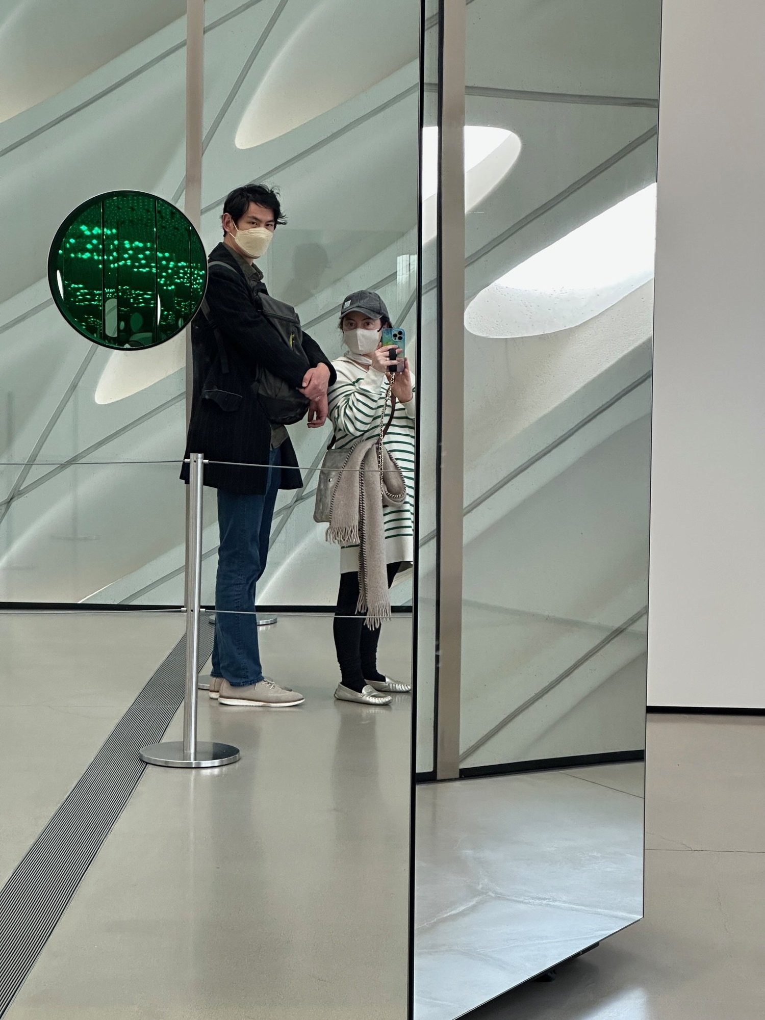 Two visitors reflected in a mirrored panel inside a bright white curved gallery space