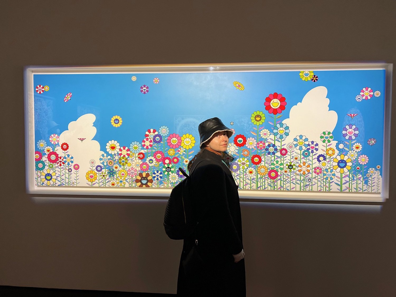 A visitor in front of a wide Takashi Murakami panel of smiling flowers under a blue sky
