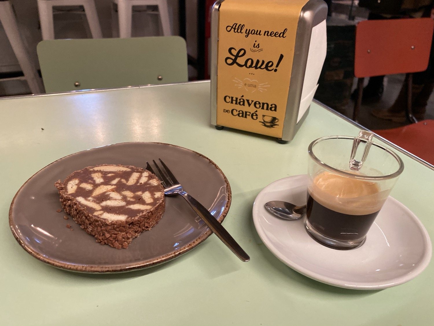 A slice of chocolate salami on a plate beside a small glass of espresso, on a pale green cafe table