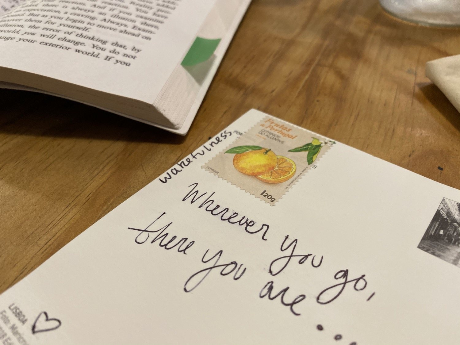 A postcard on a wooden table reading Wherever you go, there you are in handwriting, with a Portuguese orange stamp