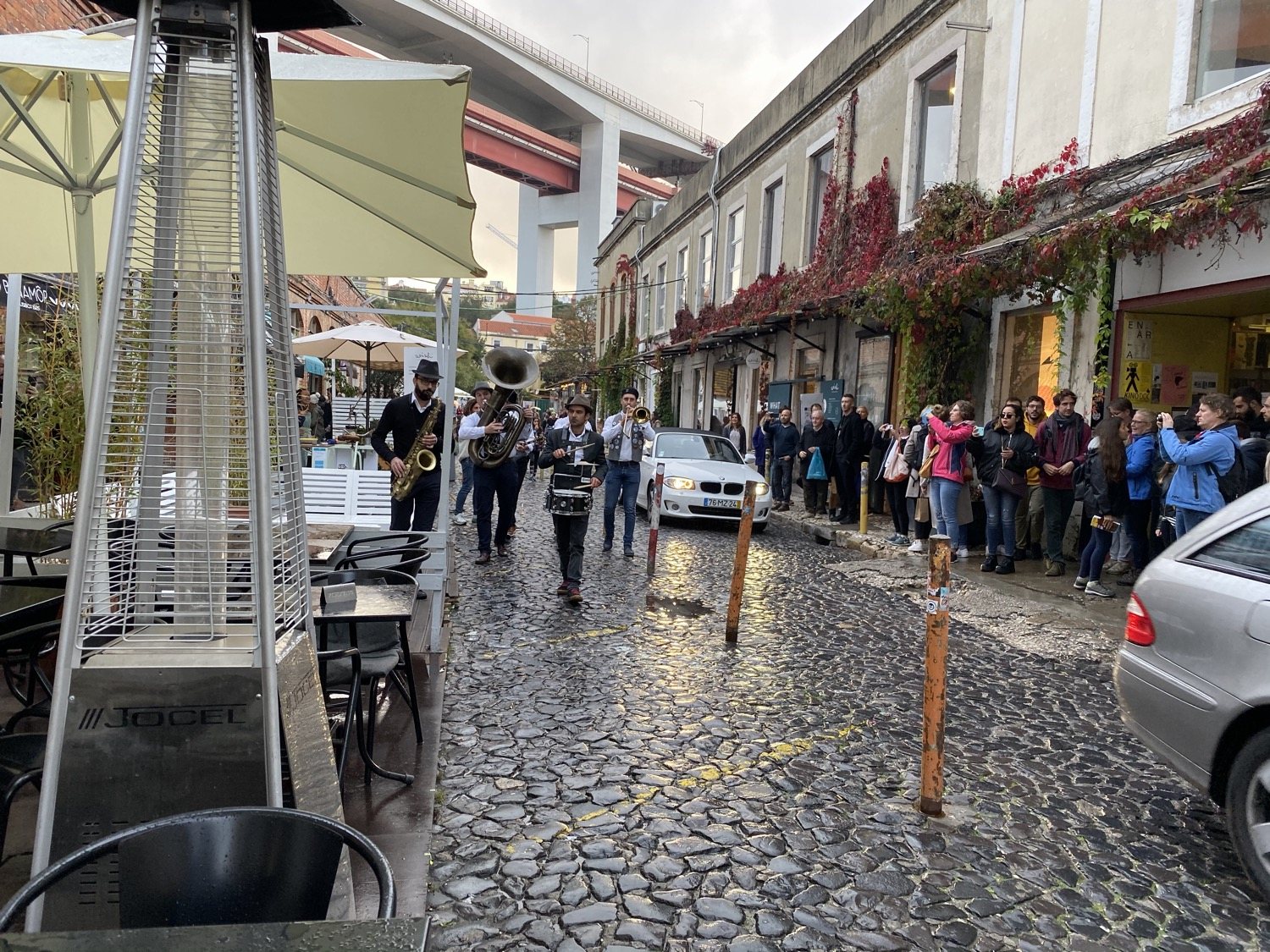 A brass band playing on the wet cobbles of LX Factory beneath the red 25 de Abril bridge, with a crowd watching from the pavement