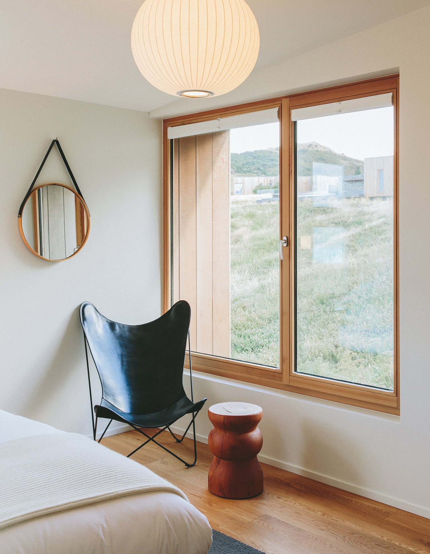 A quiet bedroom corner with a black leather butterfly chair, a round mirror on a leather strap and a window looking out to a green hillside