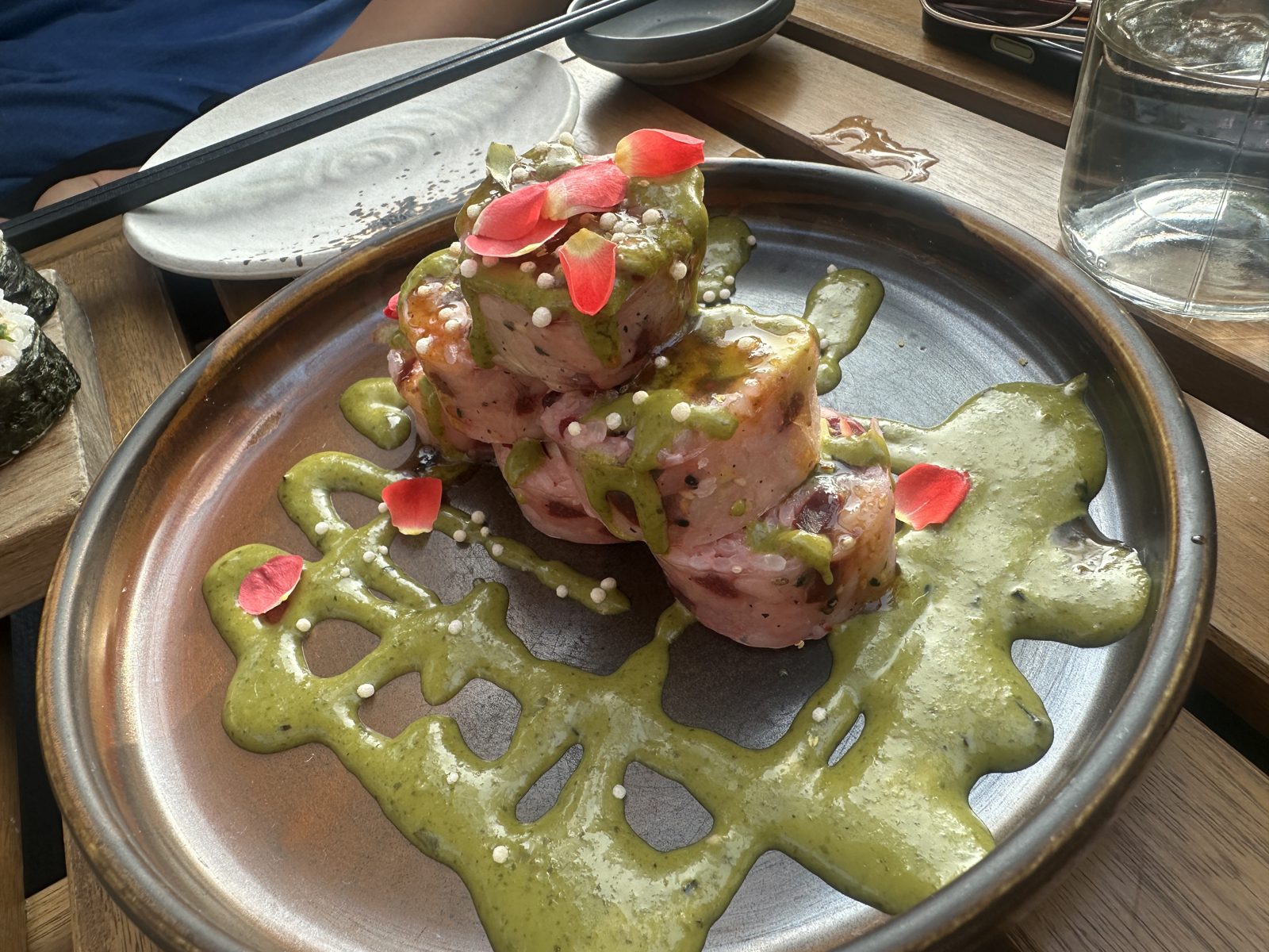 Three stacked rolls wrapped in pale pink, drizzled with a green herb sauce and scattered with small white puffed pearls and coral flower petals, on a dark ceramic plate with more of the sauce poured across it