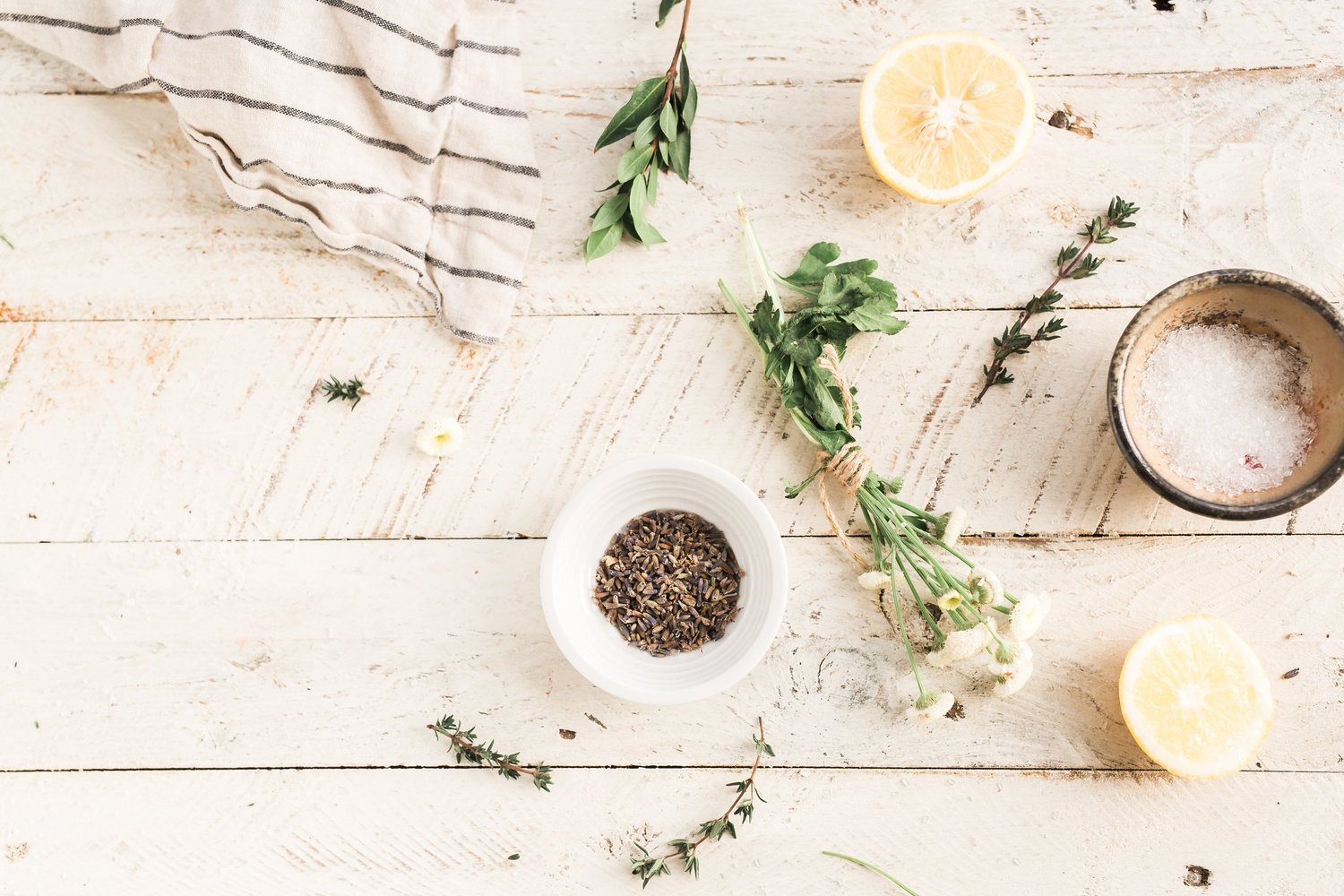 A flat lay on white painted wood with a striped linen towel, halved lemons, sprigs of thyme and small bowls of lavender and salt
