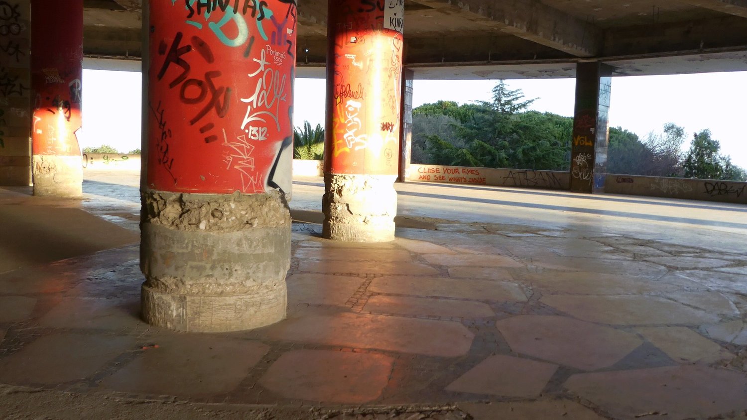 Red painted columns tagged with graffiti on an open floor, with the words close your eyes and see what is inside on the far wall