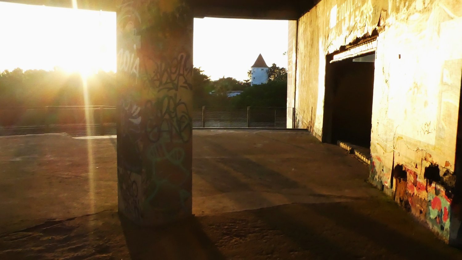 Low sun flaring across the open concrete floor, with a small white chapel tower visible through the opening