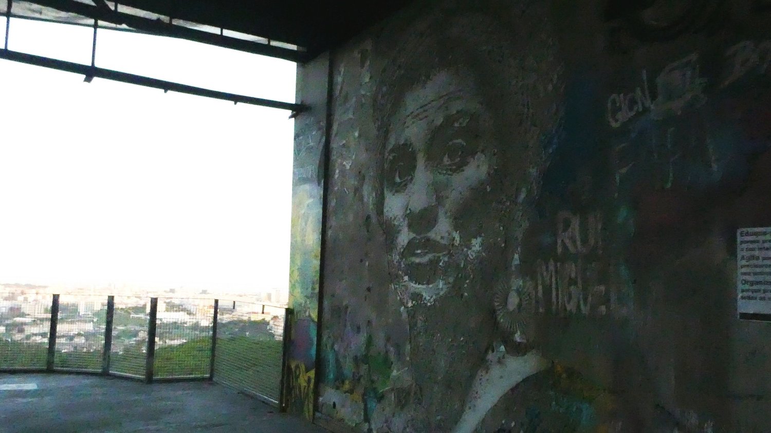A weathered painted face on the concrete wall of the viewpoint, with the Lisbon skyline through the railings