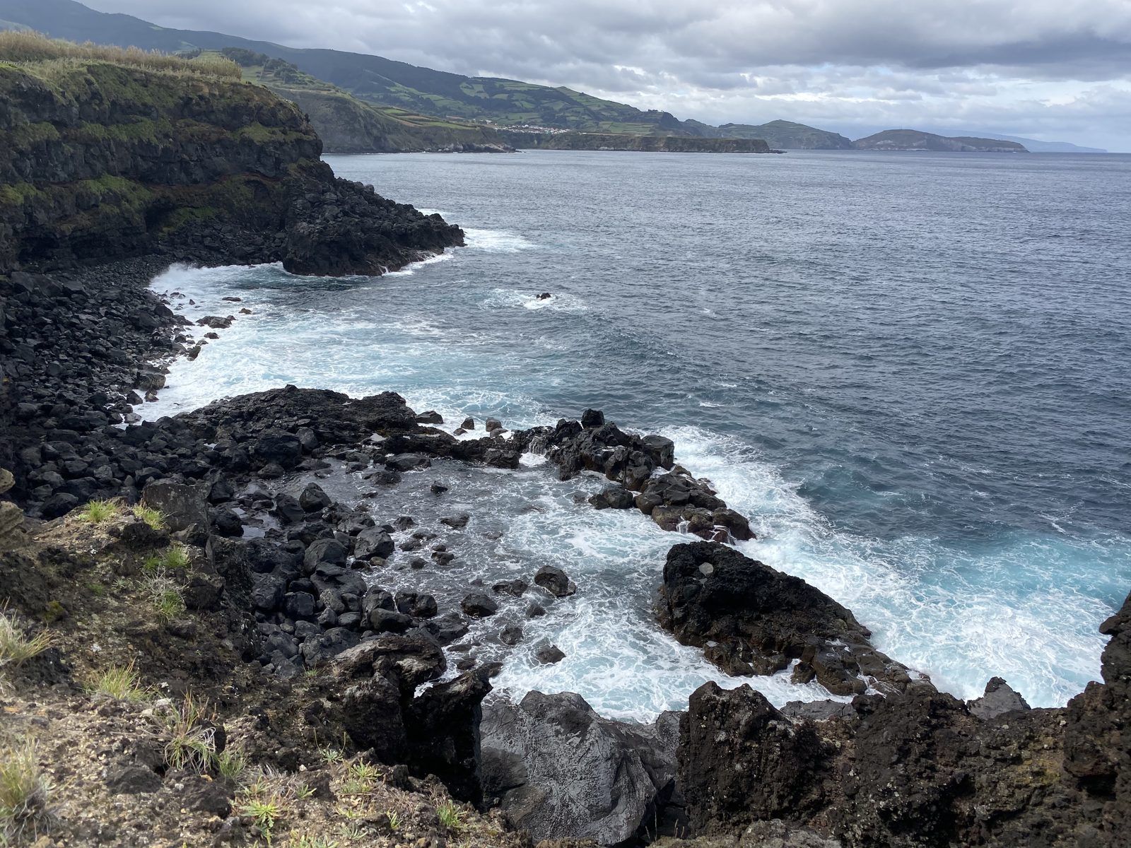 Waves breaking white against a black lava shoreline, green cliffs running away into the distance