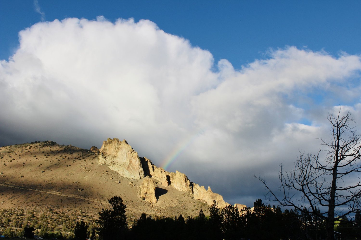 A faint rainbow falling behind the Smith Rock ridgeline under a towering white cloud