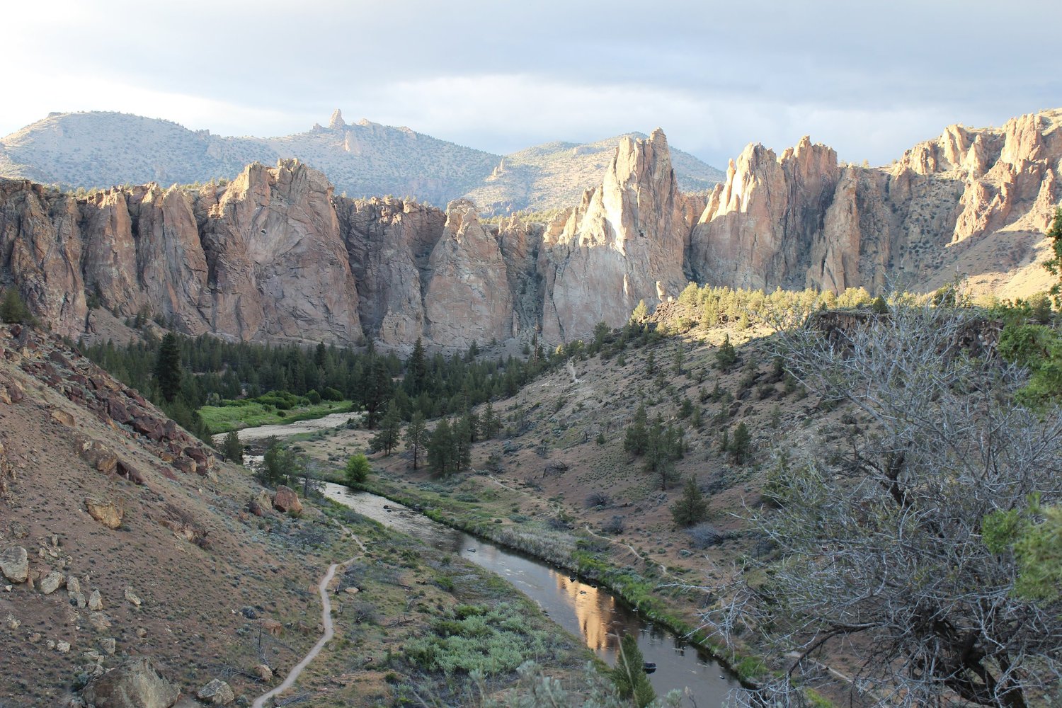 A wide view over the Smith Rock canyon at golden hour, with the Crooked River and a footpath winding below the cliffs