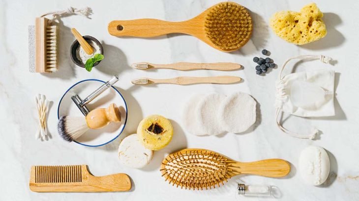 Plastic free bathroom items laid out on marble, including bamboo toothbrushes, wooden brushes, a safety razor and reusable cotton rounds
