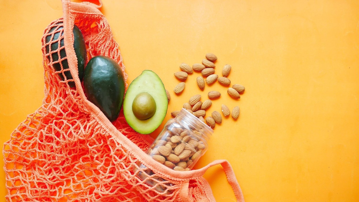Avocados and a jar of almonds spilling from an orange string shopping bag against a yellow background