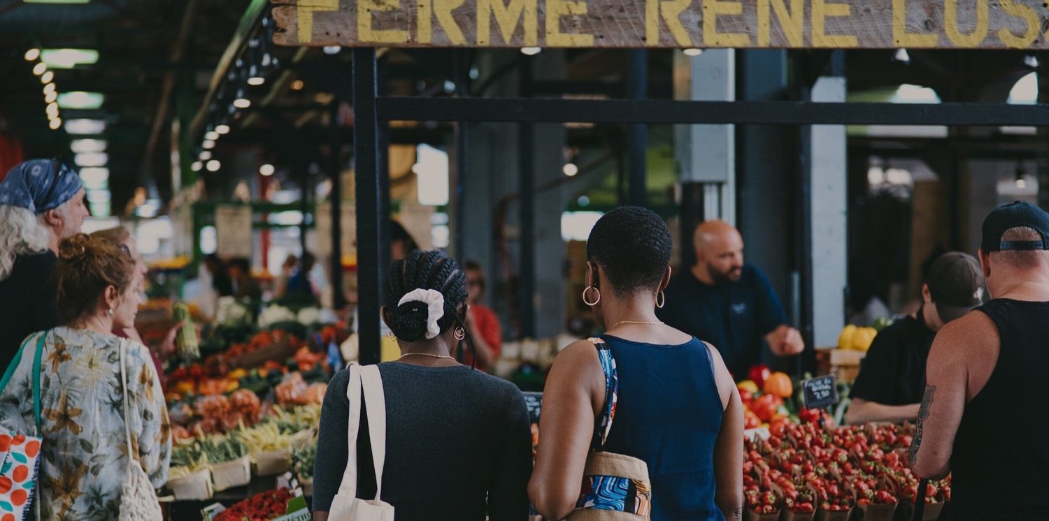 Shoppers browsing stalls piled with peppers and strawberries beneath a hand painted farm sign at a Montreal market