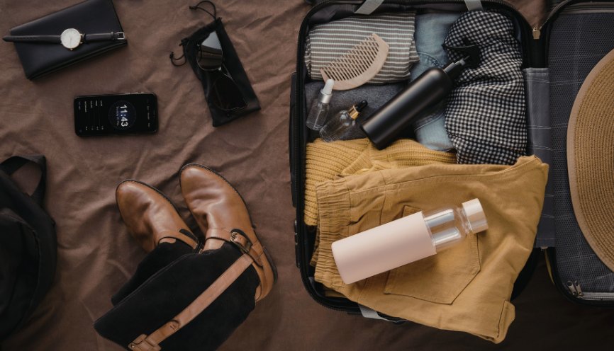 An open suitcase packed with folded clothes, a water bottle and a wooden comb, laid out beside leather boots, sunglasses, a watch and a phone on a brown blanket