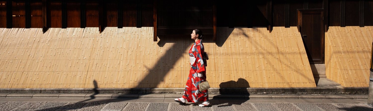 A woman in a red patterned kimono walking past a bamboo fence along a sunlit Kyoto street