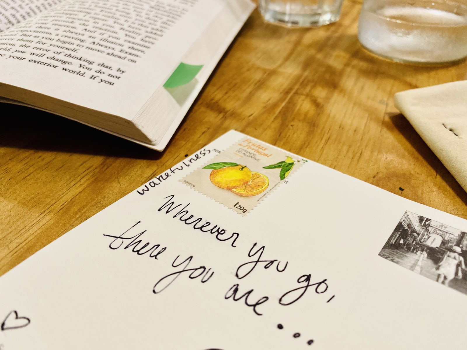 An envelope on a wooden table with Wherever you go, there you are written across it by hand, next to a Portuguese stamp of Algarve citrus and an open book