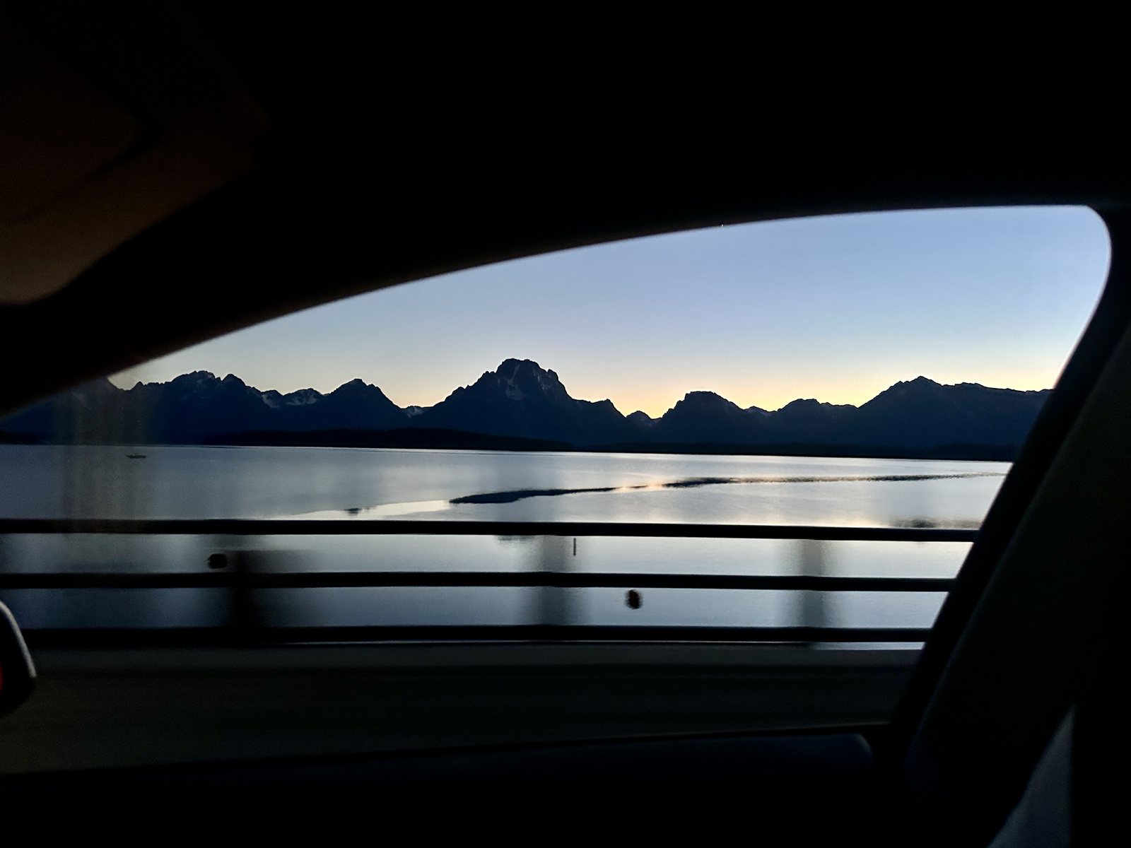 The Teton range in silhouette at dusk, seen across a lake through a car window